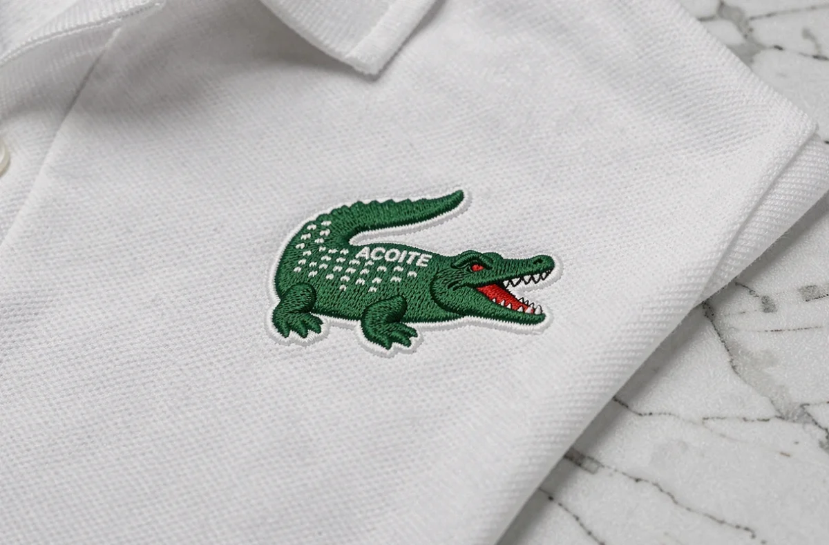 Gros plan sur le logo crocodile brodé d'un polo Lacoste blanc, montrant les détails des fils verts et la texture du tissu piqué