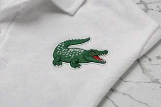 Gros plan sur le logo crocodile brodé d'un polo Lacoste blanc, montrant les détails des fils verts et la texture du tissu piqué