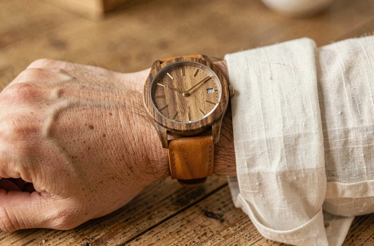 Montre en bois et cuir caramel portée au poignet d'un homme en chemise lin blanc cassé, posé sur un bureau en chêne vieilli