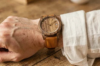 Montre en bois et cuir caramel portée au poignet d'un homme en chemise lin blanc cassé, posé sur un bureau en chêne vieilli